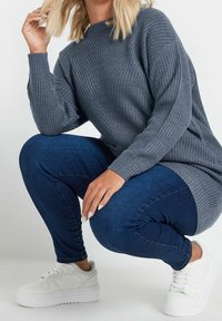 Suéter gris de punto con textura acanalada, combinado con jeans ajustados de color azul oscuro y zapatillas de plataforma blancas. Persona agachada con una mano en el cabello.