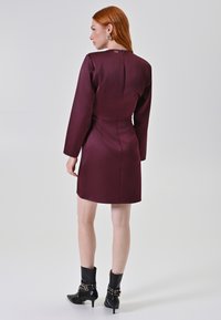 Abito lungo di colore bordeaux con maniche lunghe, vita aderente e una discreta chiusura lampo sul retro. Indossato con stivali neri alla caviglia con dettagli decorativi.