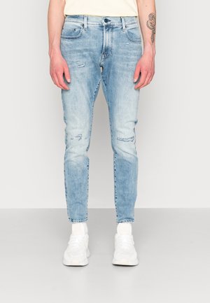 Muž v úzkých světle modrých roztrhaných džínách a bílých teniskách stojící proti jednoduchému světle šedému pozadí.