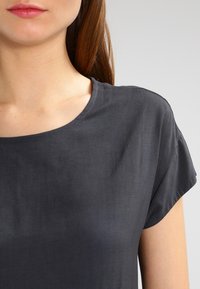 Blouse à manches courtes gris foncé en tissu lisse avec un col rond. La finition comprend des détails de couture subtils et une coupe décontractée.