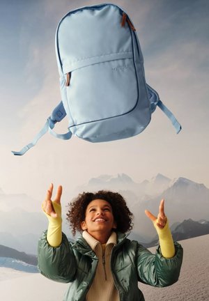 Hellblauer Rucksack mit glatter Oberfläche, ausgestattet mit einer Fronttasche und verstellbaren Trägern, über einer Person in Winterkleidung schwebend.