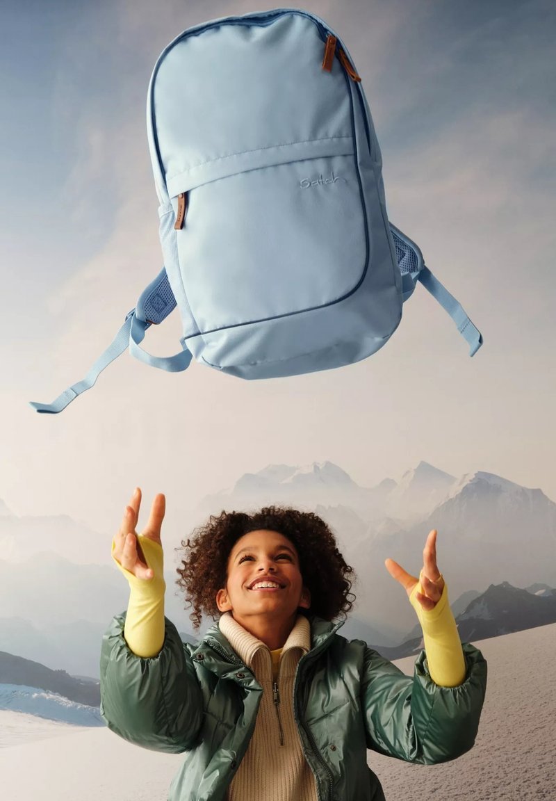 Light blue backpack with a smooth finish, featuring a front pocket and adjustable straps, suspended above a person in winter attire.