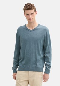 Maglione blu-grigio a maglia con scollo a V, maniche lunghe e polsini a costine. Il tessuto ha una consistenza morbida, con un piccolo logo sul petto.