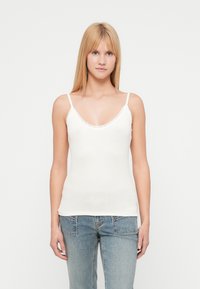 Weißes, geripptes Tanktop mit Spitzenbesatz, das dünne Träger und einen figurbetonten Schnitt hat. Kombiniert mit hellblauen Jeans.