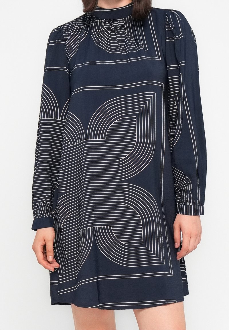 Femme portant une robe bleu marine à manches longues avec des motifs géométriques blancs et un col haut sur un fond uni.