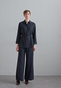 Camisa de rayas azul marino con la cintura atada, mangas largas y pantalones de pierna ancha. Tela suave con una estructura refinada. Llevada con zapatos de punta.