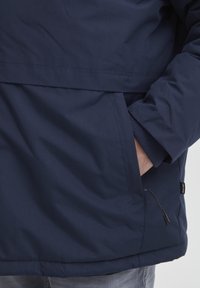 Tamsiai mėlynas lengvas švarkas su lygiu paviršiumi, priekinė kišenė, elastingi rankogaliai ir subtilus prekės ženklo ženklas ant apykaklės.