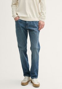 Ljusbeige tröja, blå denimjeans och vita sneakers med bruna sulor. Jeans har en standard passform och minimal detaljering. Släta texturer.