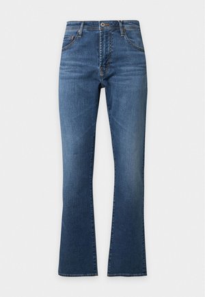 Junger Mann steht, trägt ein beiges T-Shirt mit kurzen Ärmeln, blaue Jeans, braune und beige Loafers, mit einer Hand in der Tasche vor einfarbigem Hintergrund.