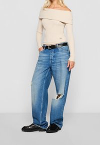 Frau trägt ein beiges Off-Shoulder-Oberteil, lockere, zerrissene blaue Jeans mit schwarzem Gürtel und schwarze Struktur-Schuhe, steht mit den Händen in den Taschen.