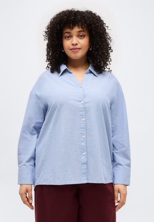 Femme aux cheveux bouclés portant une chemise boutonnée bleu clair et un pantalon rouge foncé, debout devant un fond blanc uni.
