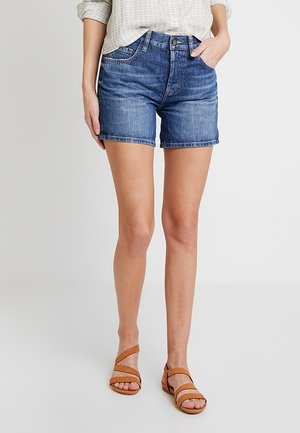 Kvinde iført blå denimshorts, lyse ternet top og brune flade sandaler med stropper, står mod en ensfarvet lys baggrund.