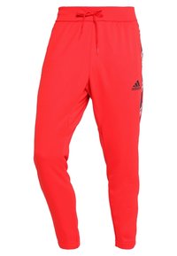 Pantalons de survêtement rouges Adidas avec taille à cordon et logo noir sur la cuisse supérieure, présentant des panneaux latéraux à motifs et des jambes fuselées.