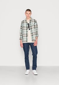 Camicia a quadri nei toni di blu chiaro, crema e nero, sopra a una maglietta grafica beige. Jeans in denim scuro e sneakers bianche completano l'outfit.