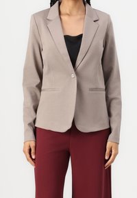 Kvinna iförd en taupe enfärgad blazer med en knapp över en svart topp och burgundy-byxor, stående framför en neutral vit bakgrund.