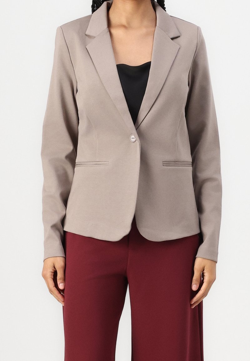 Kvinna iförd en taupe enfärgad blazer med en knapp över en svart topp och burgundy-byxor, stående framför en neutral vit bakgrund.