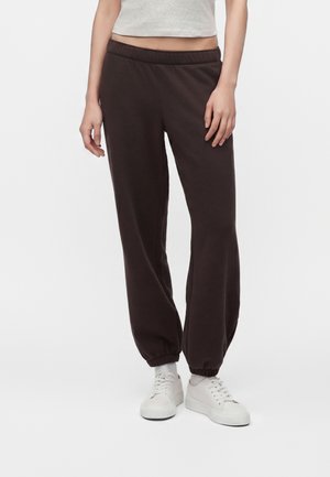 Persona che indossa pantaloni della tuta marrone scuro e scarpe da ginnastica bianche, in piedi contro uno sfondo semplice con le mani rilassate lungo i fianchi.