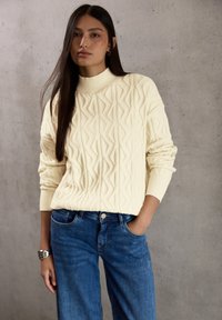 Femme aux longs cheveux noirs portant un pull crème en tricot torsadé et un jean bleu, debout avec une main dans la poche contre un mur gris.