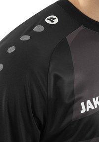 Schwarzes Sportshirt mit glatter Textur, mit grauem Akzent und reflektierenden Punkten entlang des Ärmels. Logo auf der Schulter gedruckt.