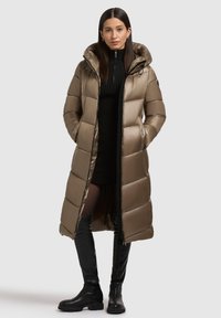 Manteau matelassé long vert olive avec capuche, présentant des sections matelassées horizontales, une fermeture éclair à l'avant et deux poches latérales. Associé à une robe pull noire et des bottes noires longues.
