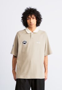 Camisa polo beige con cuello blanco, de mangas cortas, que presenta un parche con logo y texto en el pecho. Textura suave y corte relajado.