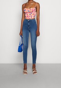 Blommig kort topp i rosa med blå accenter; högmidjade blå jeans; blå handväska; vita sandaler med remmar; släta texturer genomgående.