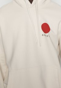 Felpa con cappuccio color crema realizzata in tessuto morbido, con una grafica centrale rossa a cerchio e testo giapponese. Include una tasca frontale e un cappuccio con cordini.