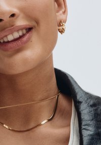 Kultaiset geometriset niittikorvakorut ja tyylikäs kultainen ketjukaulakoru paljaan ihon yllä, kehystettynä tummalla nahkatakilla. Hienovaraiset tekstuurit ja kiilto.