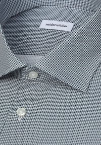 Gros plan d'une chemise pour homme à motifs avec de petites formes géométriques, des boutons blancs et une étiquette "seidensticker" visible à l'intérieur du col.