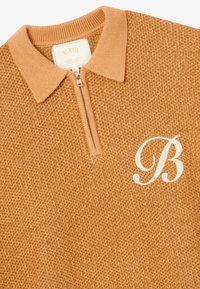 Pull en maille douce et texturée caramel à fermeture quart de zip. Avec un col côtelé et une lettre "B" brodée en blanc sur le côté gauche.