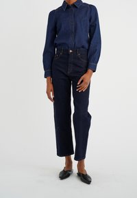 Donkerblauwe denim shirt met knoopsluiting aan de voorkant, lange mouwen en geraffineerde manchetten, gecombineerd met donkerblauwe high-waisted jeans en zwarte platte schoenen.