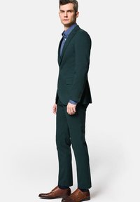 Ciemnozielony garnitur o dopasowanym kroju, z teksturowanej tkaniny, z pojedynczym zapięciem na guzik i prostymi nogawkami. Noszony na jasnoniebieskiej koszuli z ciemnymi butami.