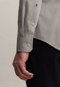 Ljust grå skjorta med en mönstrad textur, vita knappar och en svart broderad logotyp på cuffen. Cuffen har en skräddarsydd passform och sned fåll.