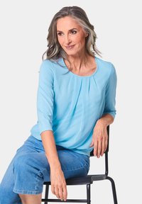Hellblaue Bluse mit Dreiviertelärmeln, Rundhalsausschnitt und gefälteltem Vorderteil, kombiniert mit hellblauen, kurzen Jeans, sitzt auf einem schwarzen Stuhl.