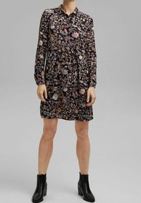 Blommönstrad klänning med långa ärmar, svart botten, krage och rynkad midjedetalj. Tillverkad av lättviktsmaterial; kombinerad med svarta ankelboots.