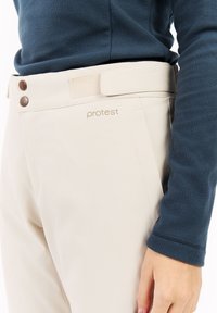 Crème kleurige broek met een gladde textuur, voorzien van een tailleband met twee bruine knopen en een logo aan de zijkant. Vergezeld door een blauwe fleece top.