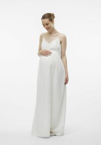 Robe de maternité longue blanche avec un corsage en dentelle, des bretelles fines, une texture fluide et une taille empire, créant une silhouette douce.