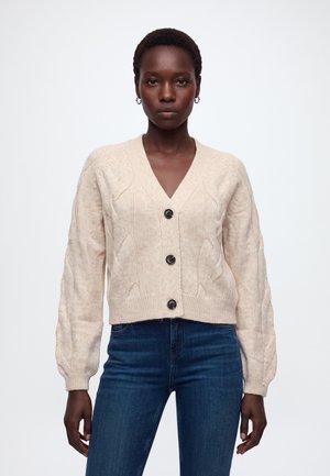 Femme portant un cardigan beige à boutons en maille torsadée et un jean bleu, debout devant un fond blanc uni.