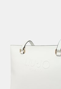 Borsa tote bianca con logo impresso "LIU·JO MILANO", dotata di accessori dorati, texture liscia e due manici superiori.