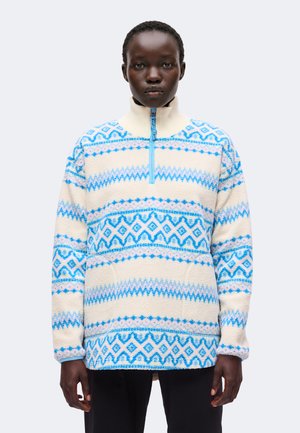 Personne portant un pull polaire crème et bleu à motif avec un col à demi-fermeture éclair, debout devant un fond clair uni.