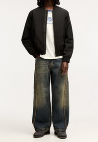 Zwarte bomberjack, witte grafische T-shirt, wijde, vervaagde blauwe jeans met oranje stitching, en zwarte sneakers. Ontspannen pasvorm en casual stijl.