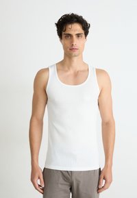 Weißes, geripptes Tanktop mit breiten Trägern, im klassischen Schnitt. Getragen mit hellgrauen Hosen vor einem neutralen Hintergrund.
