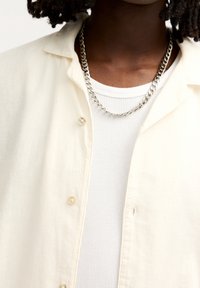 Lichtbeige overhemd met knopen over een wit geribd tanktopje, met een zilveren ketting zichtbaar bij de hals. Zachte stoffen textuur, casual ontwerp.