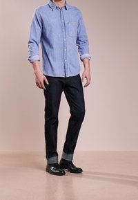 Ljusblå skjorta med uppvikta ärmar, mörka slim-fitjeans med manchetter och svarta ankelboots, mot en neutral bakgrund.