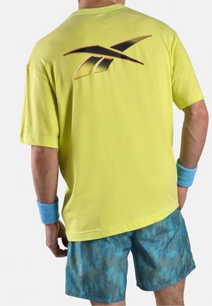 Mann trägt gelbes Sport-T-Shirt mit schwarzem und orangefarbenem Reebok-Logo auf dem Rücken, blaue Camouflage-Shorts und blaue Handgelenkbandagen, steht mit dem Rücken zur Kamera.