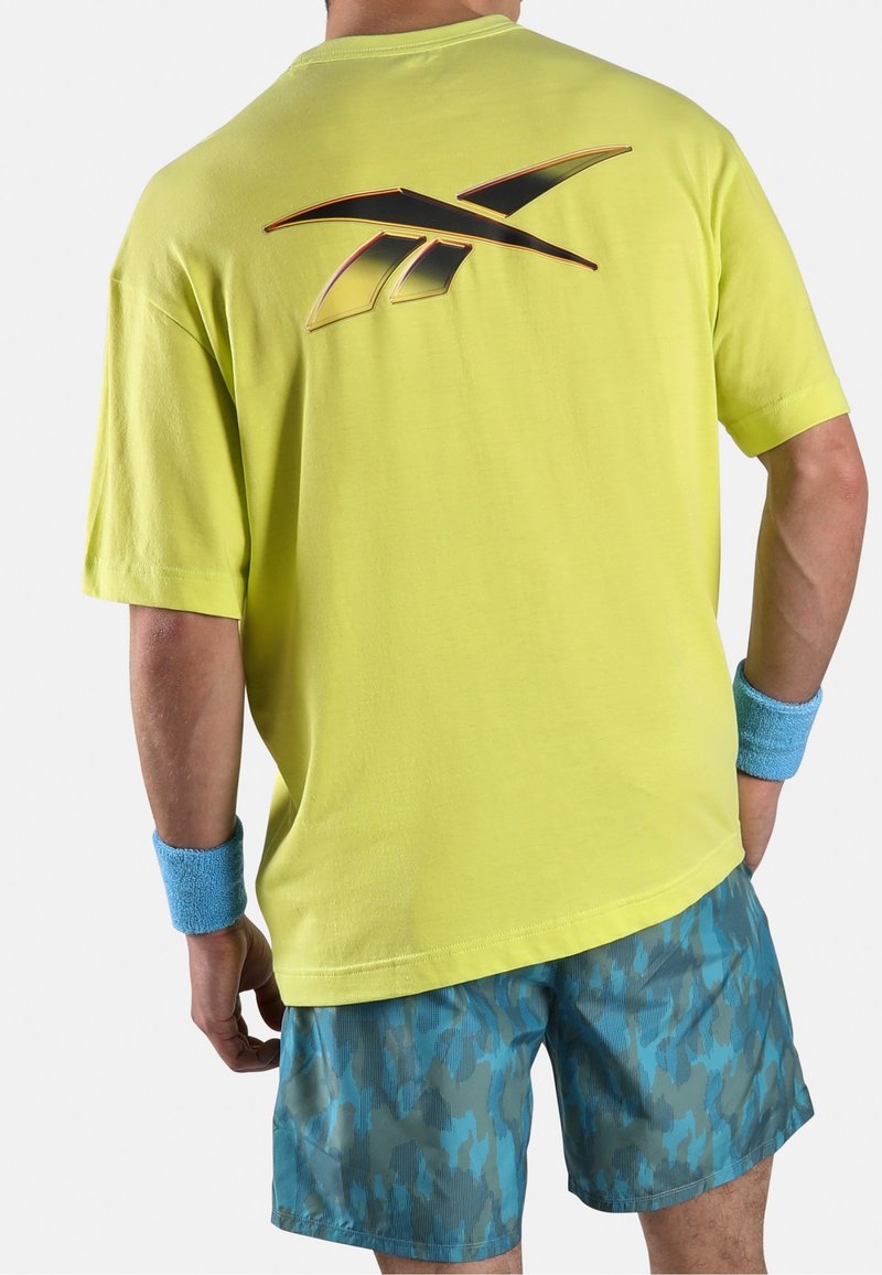 Man wearing yellow athletic T-shirt with black and orange Reebok logo on back, blue camouflage shorts, and blue wristbands standing with back to camera.
