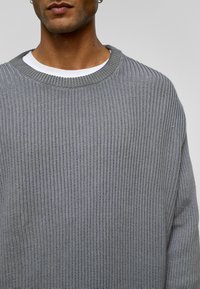 Grijze gebreide trui met een brede, ribbelstructuur, ronde halslijn en verlaagde schouders. Gelaagd over een wit shirt.