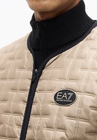 Κοντινό πλάνο ενός ατόμου που φοράει μπεζ καπιτονέ μπουφάν με μαύρο φερμουάρ και σήμα λογότυπου EA7 Emporio Armani στο στήθος.