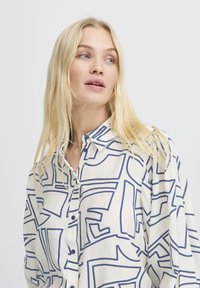 Camicia button-up in crema con audaci motivi geometrici navy. Presenta colletto, maniche lunghe e una vestibilità rilassata. Il tessuto appare morbido.