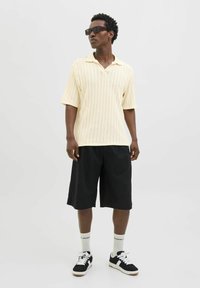 Polo shirt a manica corta in maglia crema con colletto aperto, abbinato a pantaloni corti neri al ginocchio. Indossato con sneakers bianche e nere e occhiali da sole.